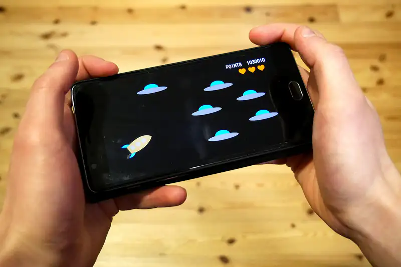 Two hands holding a smartphone playing a space-shooter game on a wooden table